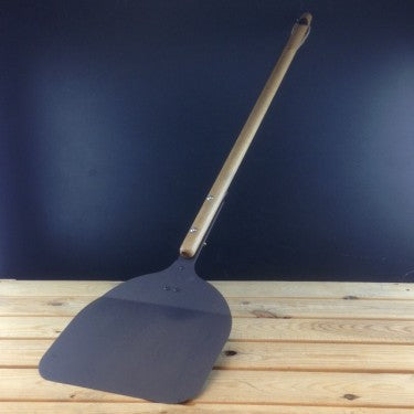Black iron pizza peel with long oak handle resting on outdoor oven